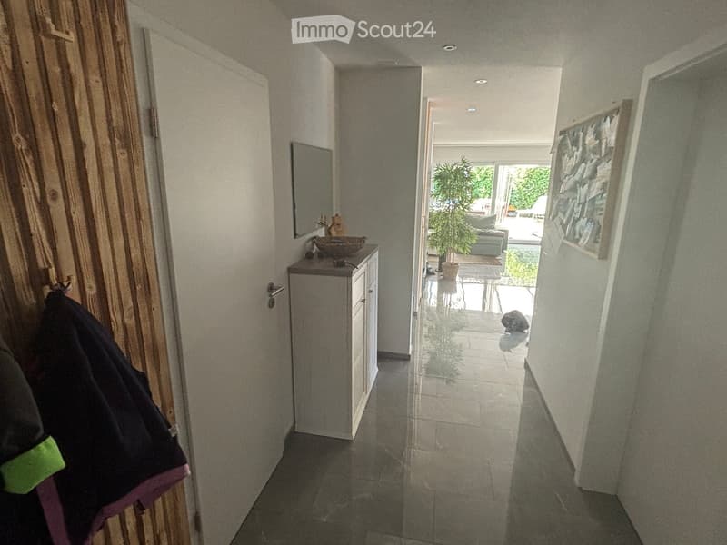 Hallway with tiled floor, white walls, small cabinet with a bowl on top, coat hanging on the left, and a view into a living area with a plant and sofa.