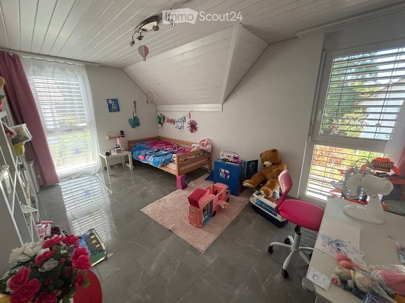 A cozy children's bedroom with a bed, chair, table, toys, and a fan. The room features a slanted ceiling, large windows, and a colorful, playful decor.