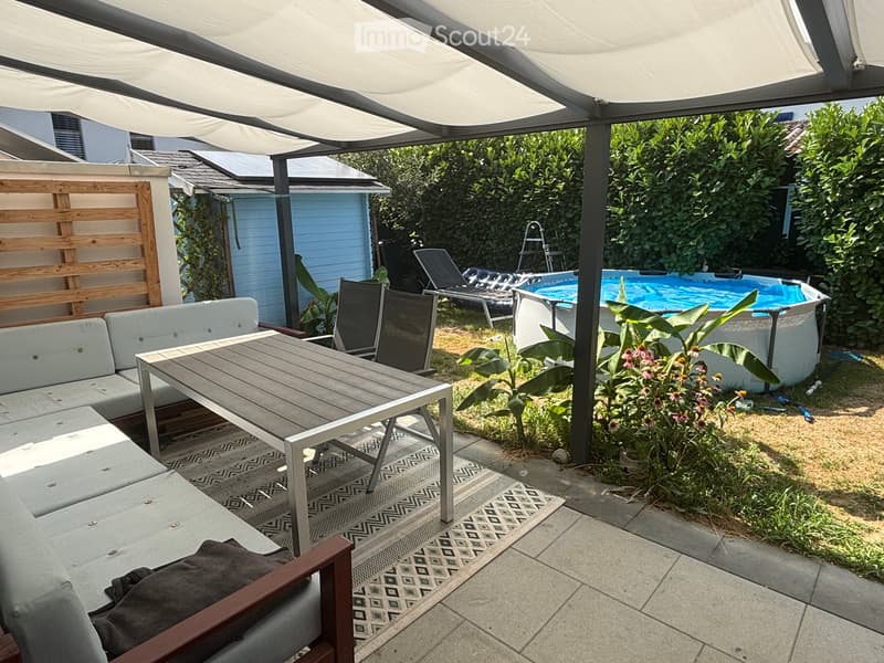 Outdoor patio with a grey sofa, wooden table, chairs, and a swimming pool surrounded by greenery and a wooden fence.