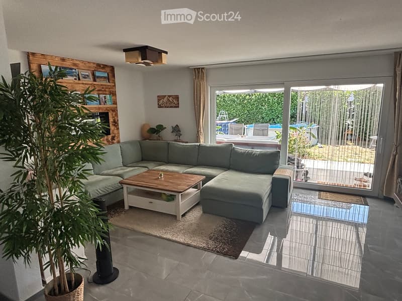 Modern living room with a grey sectional sofa, wooden coffee table, large potted plant, and a shelf with decorative items. Large sliding glass doors open to a patio with outdoor furniture and a swimming pool visible.