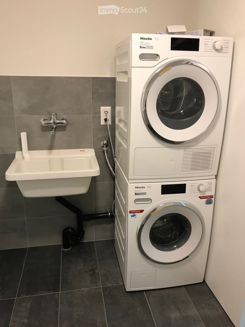 A small bathroom with a white washing machine, a white sink with a chrome faucet, and grey tiled walls and floor.