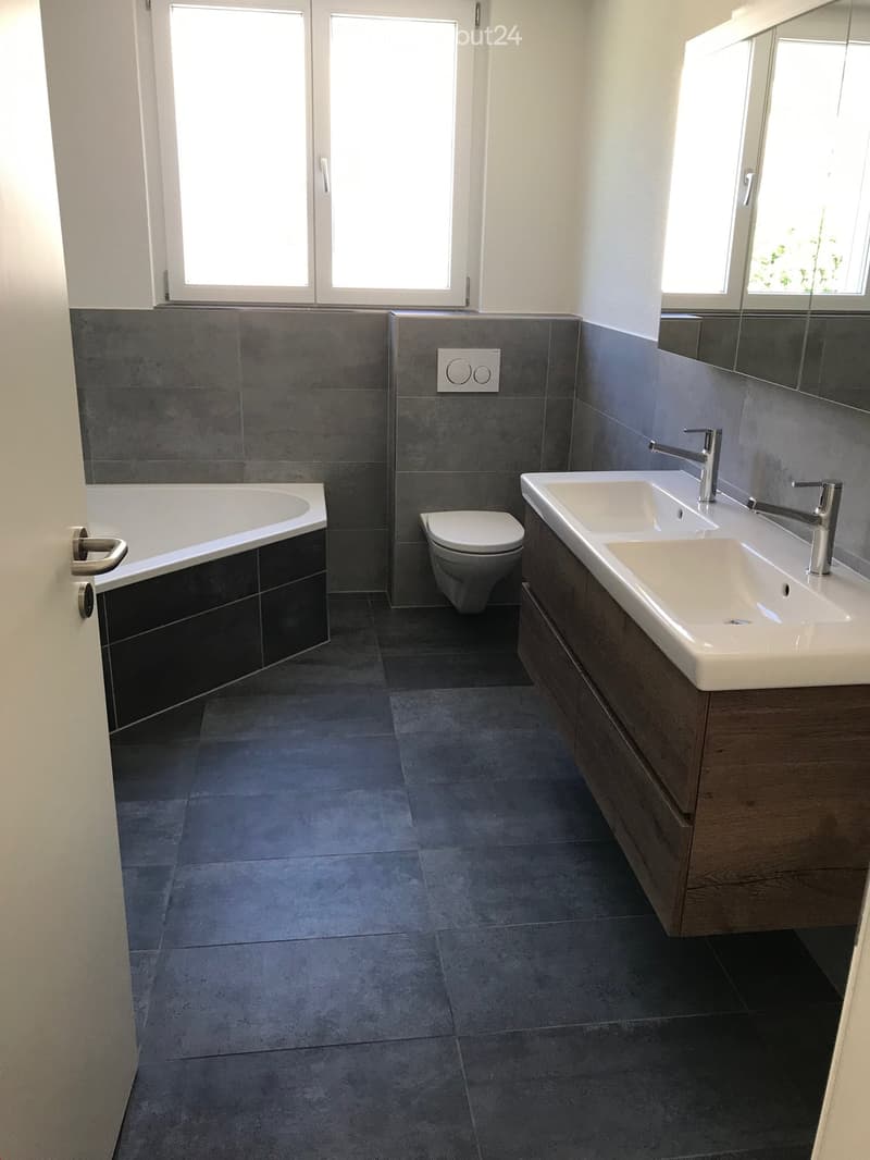 Modern bathroom with dark grey tiles, a bathtub, a toilet, a double sink with wooden cabinet, and a large mirror.