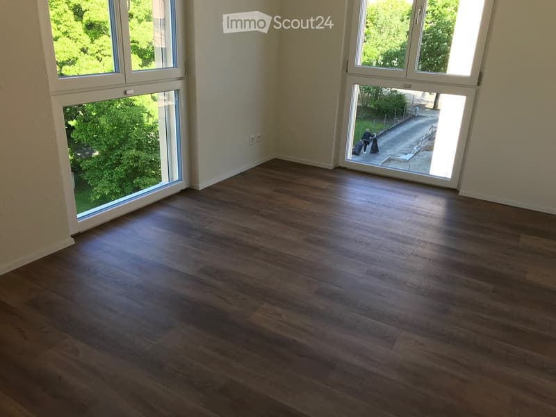 Empty room with wooden flooring and large windows offering a view of greenery outside. The space is unfurnished and appears to be a living or sitting area.