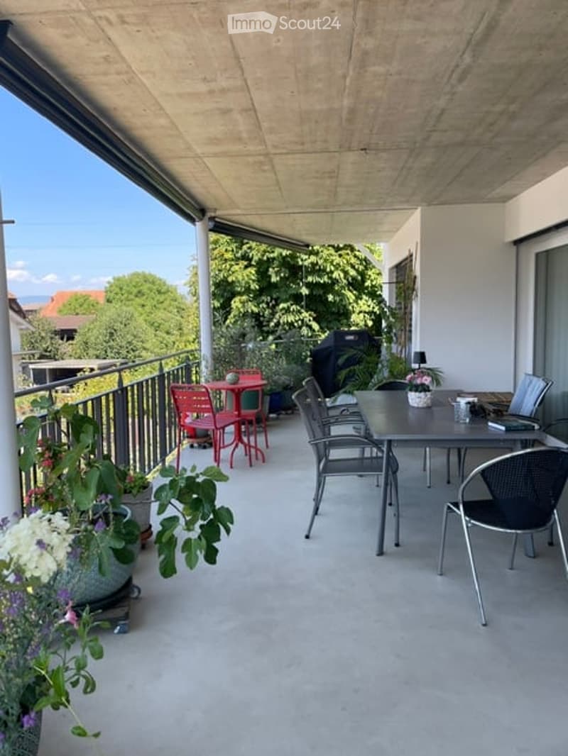 Modern balcony with concrete floor, metal railing, and outdoor furniture including a dining table, chairs, and potted plants.