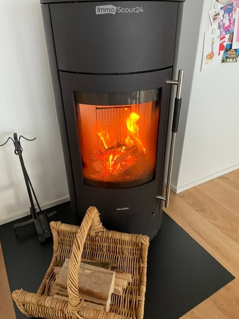 Modern wood-burning stove with visible flames, placed on a black mat, accompanied by a woven wood basket filled with logs.