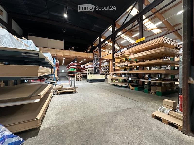 A spacious warehouse with high ceilings, metal beams, and exposed wooden supports. Shelves are stocked with wooden planks and materials, and a forklift is visible in the center.