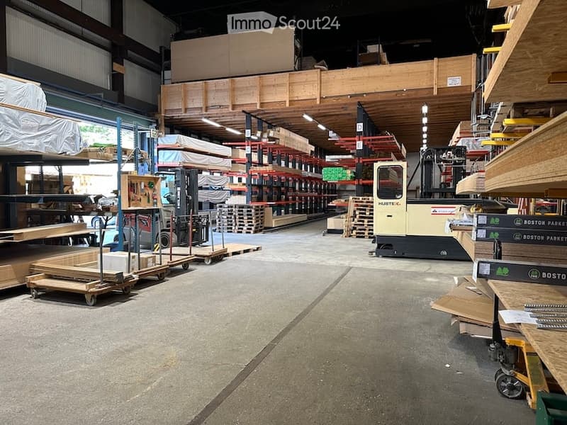 Warehouse interior with high ceilings, metal shelving units, and various construction materials including wooden planks, pallets, and machinery.
