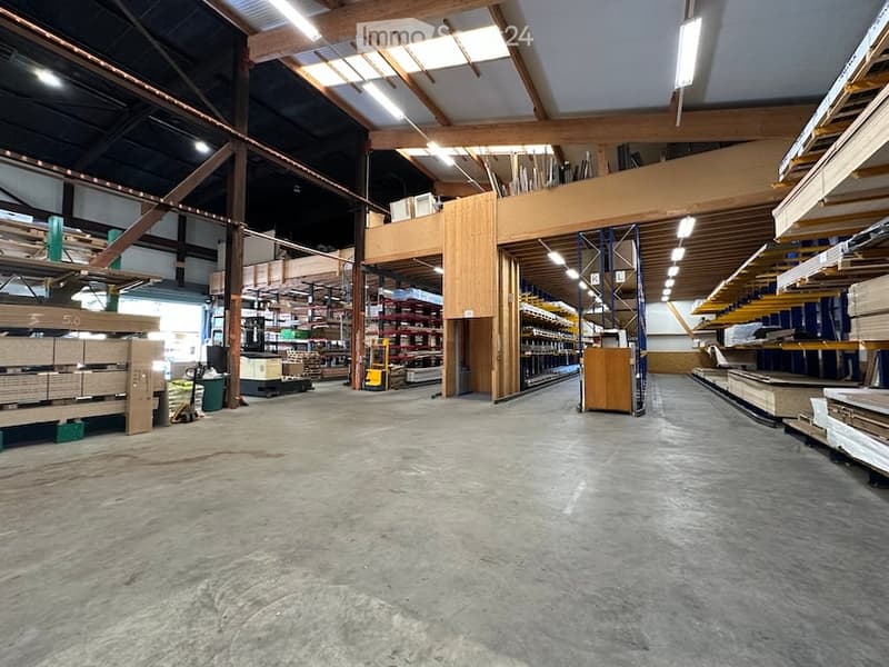 Large industrial-style warehouse with high ceilings, exposed wooden beams, and metal shelving units. The space is clean and organized, with various materials and equipment stored on shelves and racks.