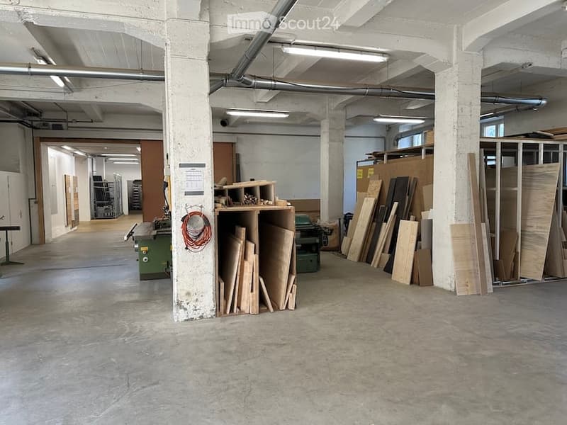Large, open industrial-style workshop or storage space with concrete floors, exposed white columns, and visible pipes. Shelves hold various wooden planks and materials, with a workbench and tools visible in the background.