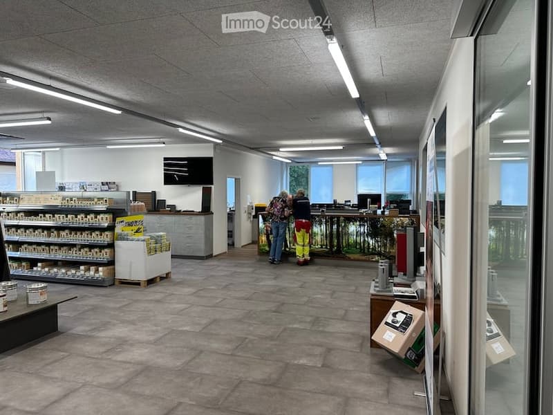 A spacious, well-lit room with a concrete floor and white walls. Shelves hold various items, a counter is visible, and a large screen is mounted on the wall. A table and chairs are present, and the room has large windows allowing natural light to fill the