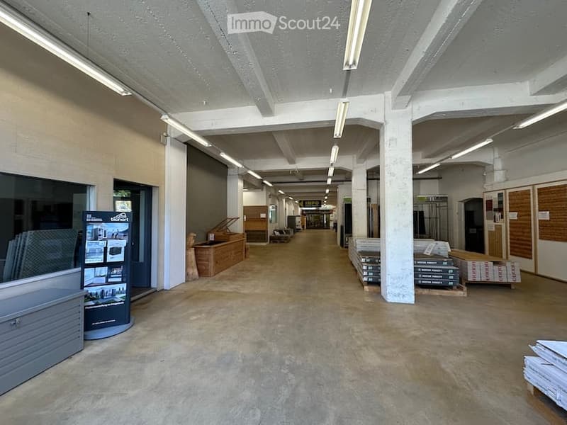 Empty industrial-style corridor with exposed concrete floors, white painted concrete pillars, and fluorescent lighting. No furniture or equipment visible.