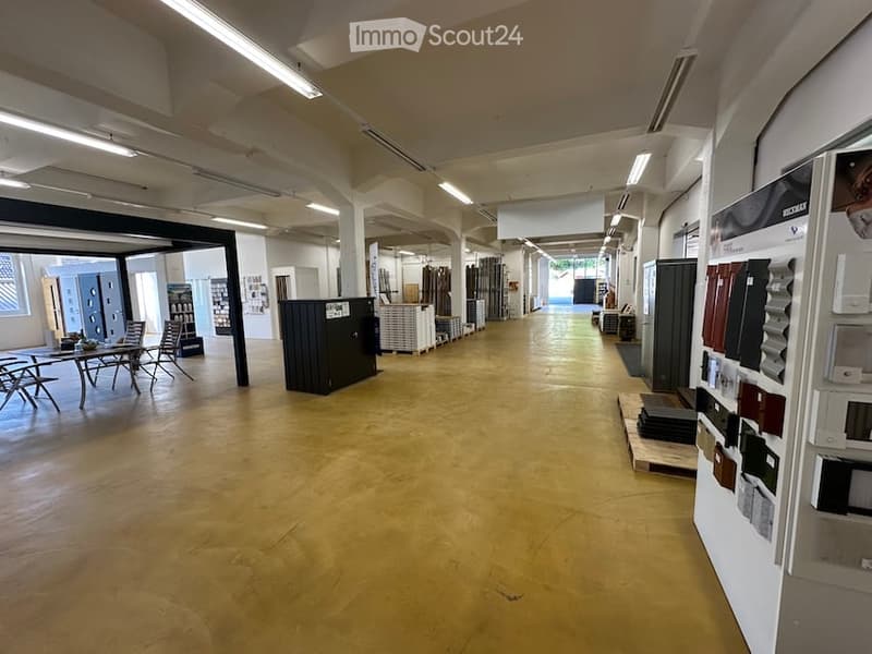 Large, open showroom space with polished wooden flooring, exposed white beams, and display areas for tiles and materials. Minimal furniture visible.