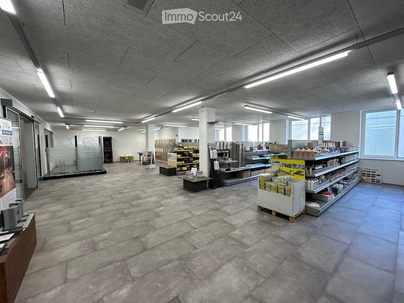 A spacious, well-lit retail or showroom space with exposed concrete ceilings, large windows, and a polished stone tile floor. Shelves and display units hold various products, with a counter and seating area visible.