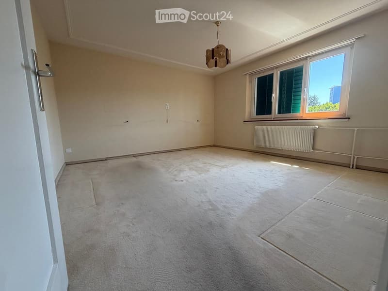 Empty room with beige walls, carpeted floor, large window with view, radiator, and chandelier.