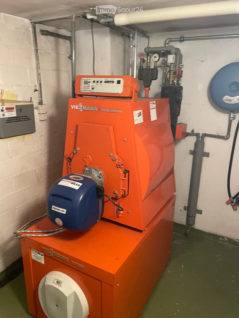 A modern orange boiler unit with a control panel, pump, and various pipes and valves installed in a utility room.