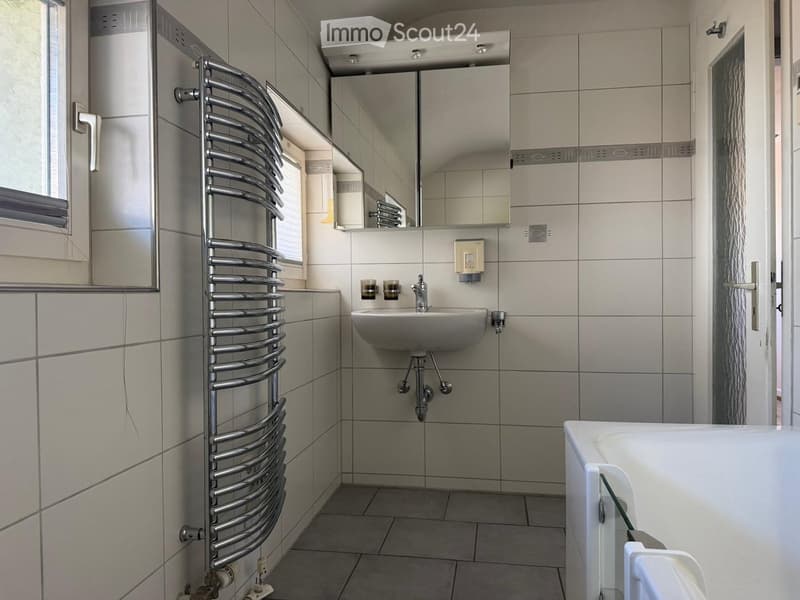 Modern bathroom with a white sink, chrome faucet, bathtub, radiator, and mirror. Light gray tiles and a window provide natural light.