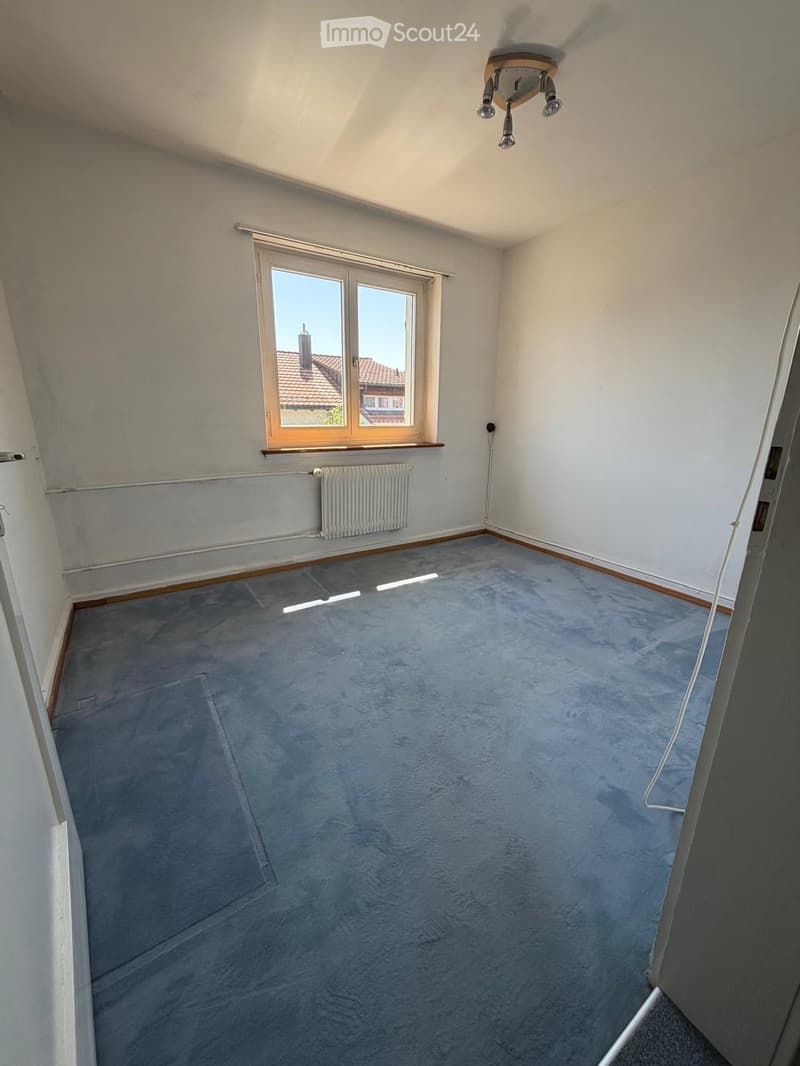 Empty room with grey carpet, white walls, large window with wooden frame, radiator, and ceiling light fixture.