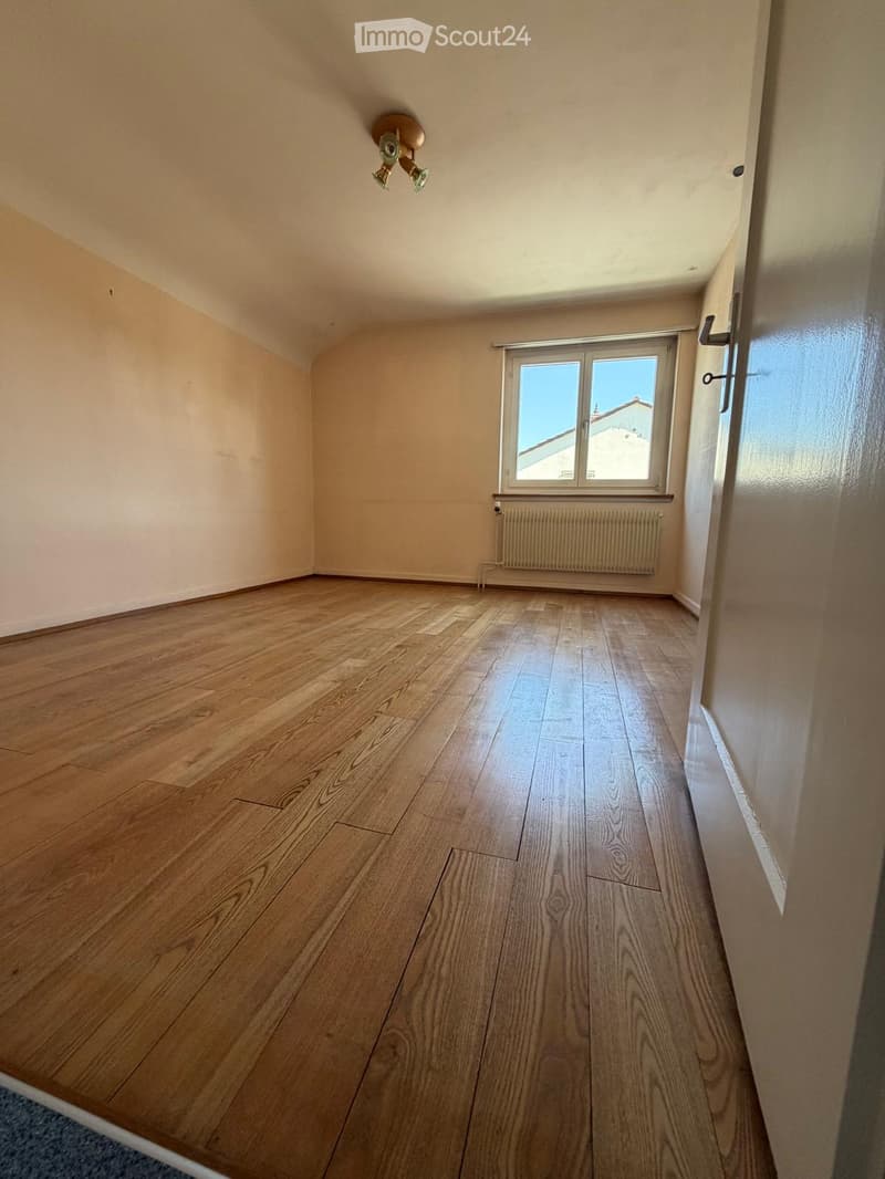Empty room with wooden flooring, white walls, a window with a view, and a radiator below the window.