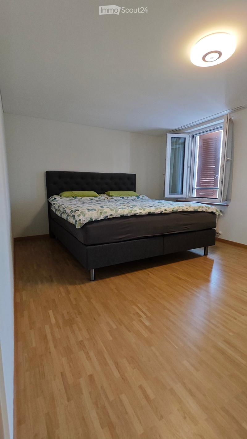 Bedroom with wooden flooring, a bed with a dark headboard and green pillows, and a large window with blinds.
