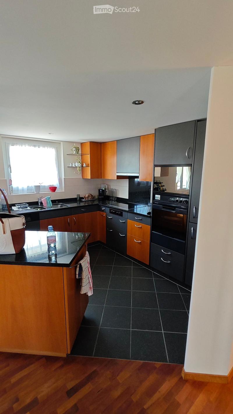 Modern kitchen with wooden and grey cabinets, black countertops, and stainless steel appliances. Features include a sink, oven, refrigerator, coffee maker, and a bottle on the counter.