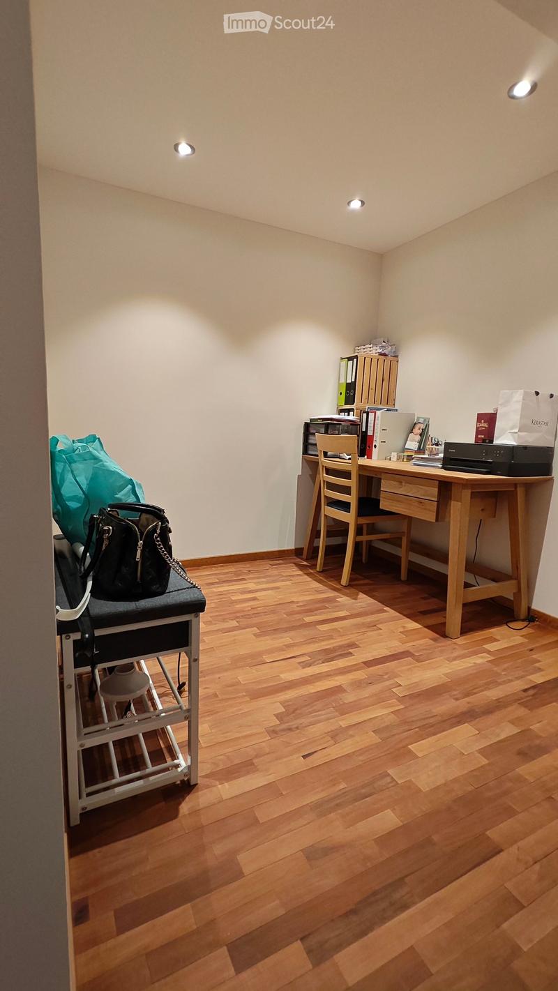 A small, well-lit room with wooden flooring and white walls. It features a wooden desk with a chair, a printer, and various office supplies. A handbag is placed on a small stool near the entrance.