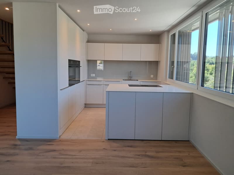 Modern kitchen with white cabinets, central island, and large windows offering a view of greenery.