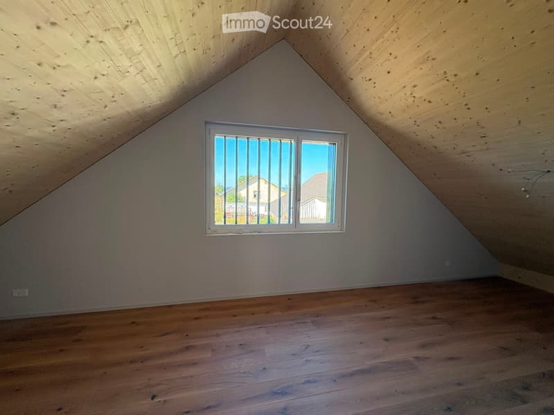 Empty attic room with wooden floors, slanted wooden ceiling, and a single window showing a view of neighboring houses.