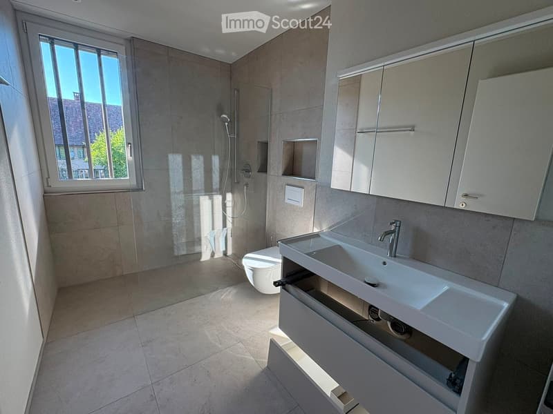 Modern bathroom with a large window, white sink, toilet, mirrored cabinet, and a glass-enclosed shower area.