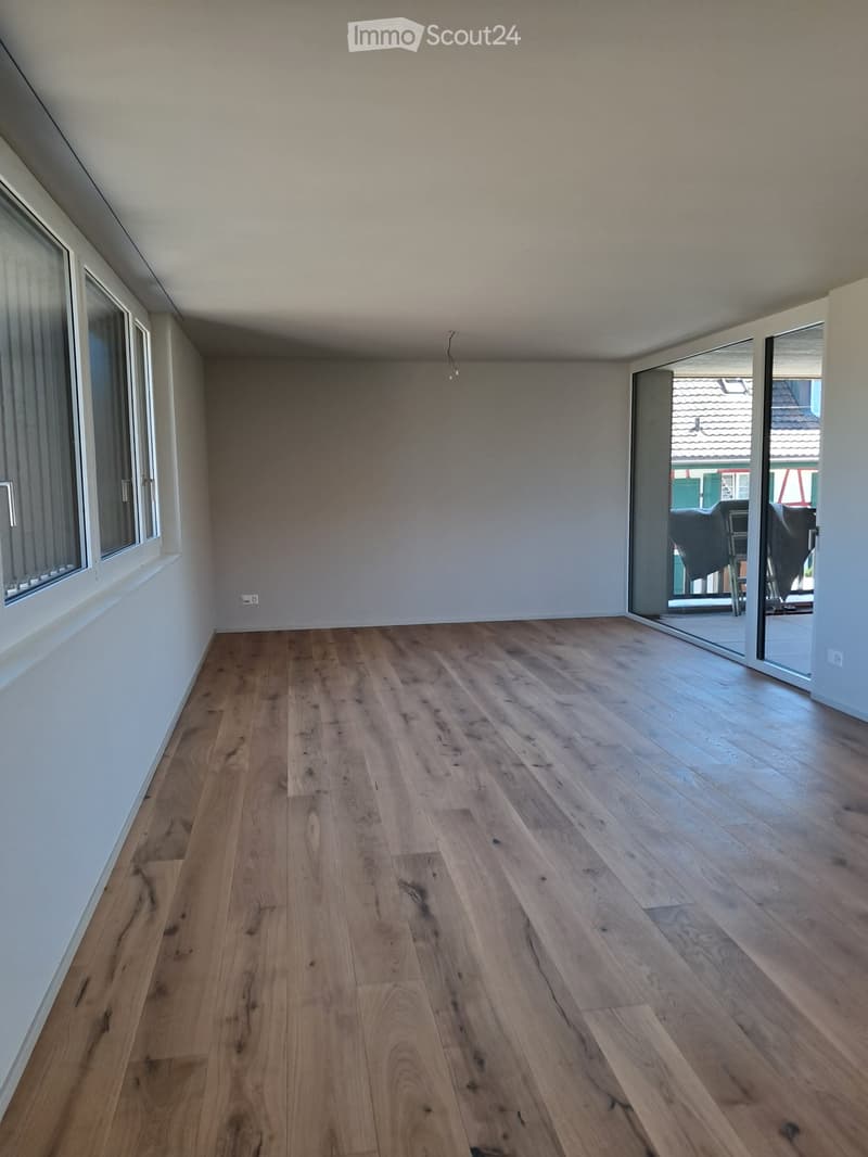 Empty room with wooden flooring and large windows, featuring a sliding glass door leading to an outdoor area.