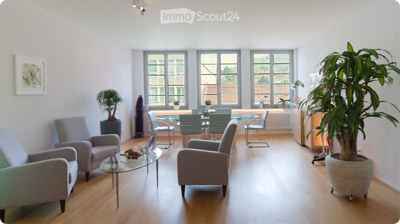 Bright living room with light wooden flooring, large windows, grey sofa, armchairs, coffee table, dining area with glass table and chairs, and several potted plants.