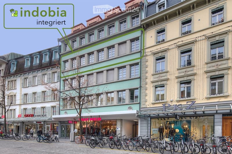 A multi-story building with a light green facade and multiple windows, located in an urban area with bicycles parked in front and storefronts visible at the ground level.