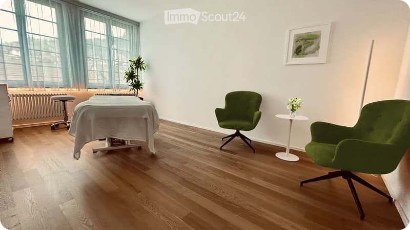 A well-lit room with wooden flooring, a massage table covered with a white sheet, two green chairs, a small round table with a vase of flowers, and a potted plant near the window.