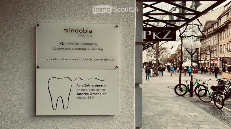 A signboard for a dental practice and massage services is mounted on a building wall, with a busy urban street scene in the background featuring pedestrians, bicycles, and storefronts.