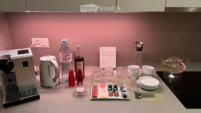 A modern kitchen counter with a coffee machine, kettle, teapot, cups, glasses, bottles, and a tray with tea bags and sugar.