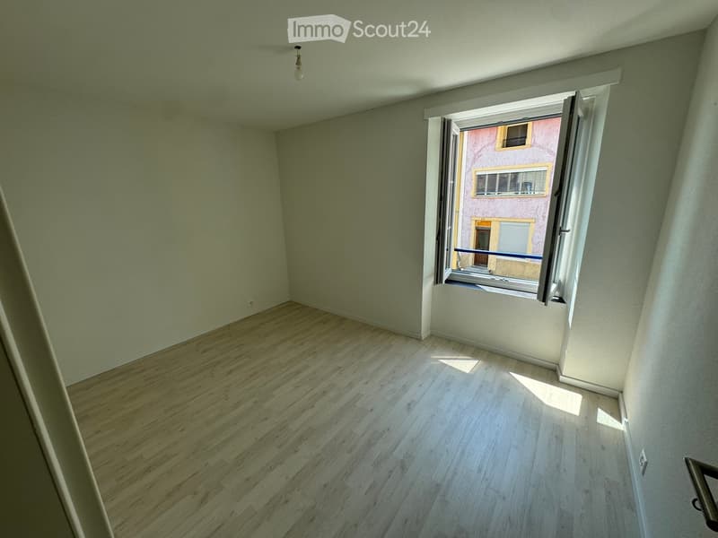 Empty room with wooden flooring and white walls, featuring an open window with a view of a neighboring building.