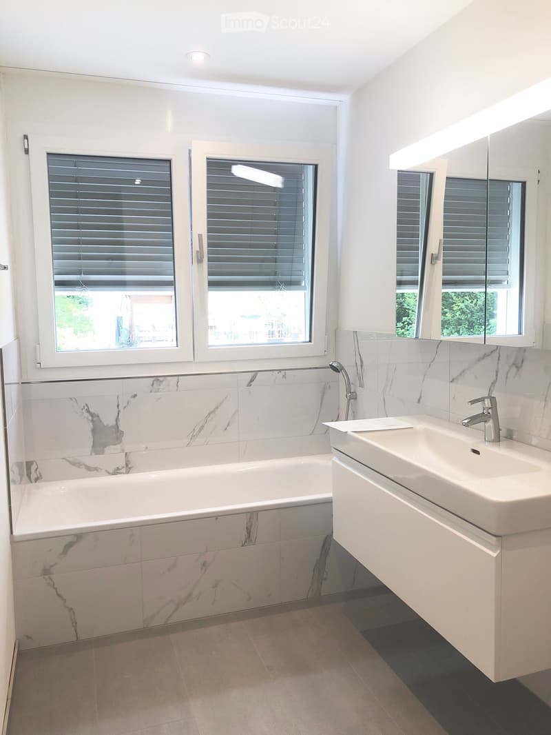Modern bathroom with a white bathtub, marble tiles, a white sink with a modern faucet, and a large mirror above the sink. The room features large windows with blinds and a sleek, contemporary design.