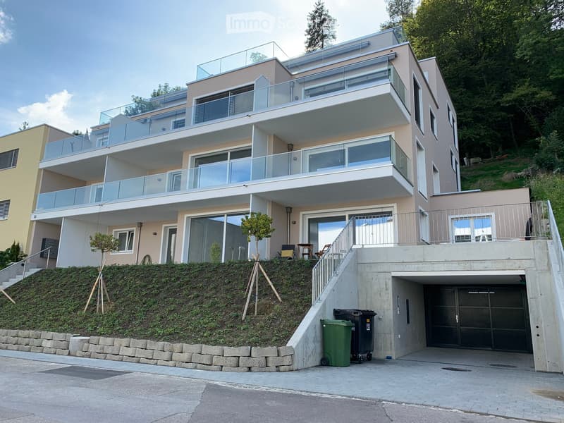 Modern two-story house with large glass balconies, raised ground floor, and garage. The house features a beige facade, multiple large windows, and a landscaped hillside entrance.