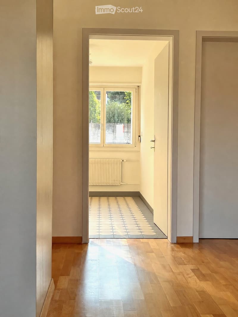 A well-lit hallway with wooden flooring, leading to a room with a tiled floor and a radiator. The walls are painted in a light color, and the space is clean and minimalistic.