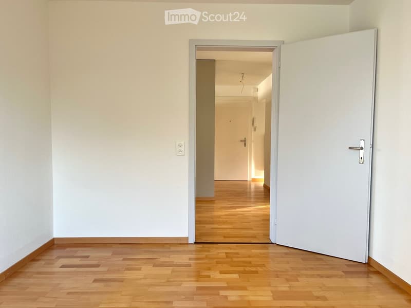Empty hallway with light wooden flooring and white walls, featuring an open door leading to another room.