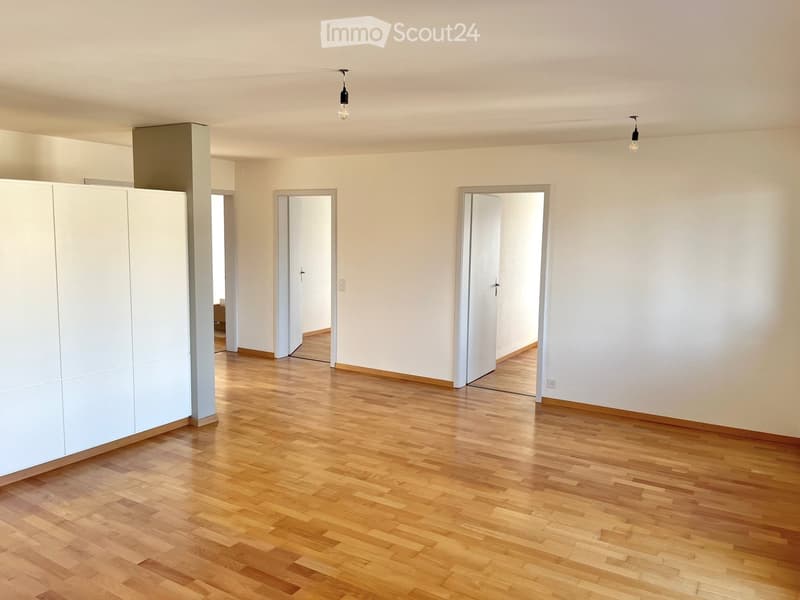 Empty room with light wooden flooring, white walls, and several open doors leading to other spaces.