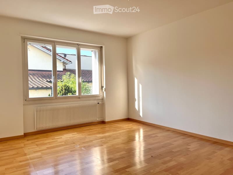 Empty room with wooden flooring, white walls, large window with view of neighboring building, radiator below window.
