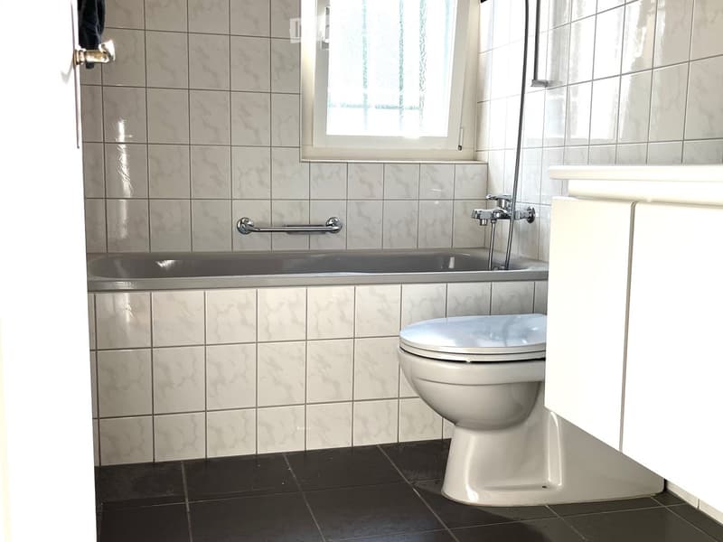 Bathroom with a toilet, bathtub, and a handrail, featuring tiled walls and a window.