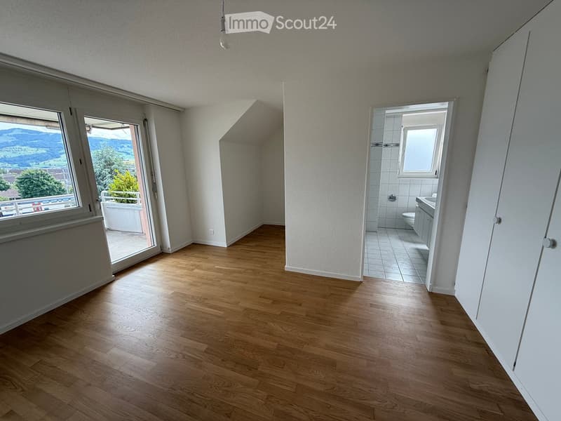 Empty room with wooden flooring, white walls, and large windows offering a scenic view. A bathroom is visible through an open doorway.
