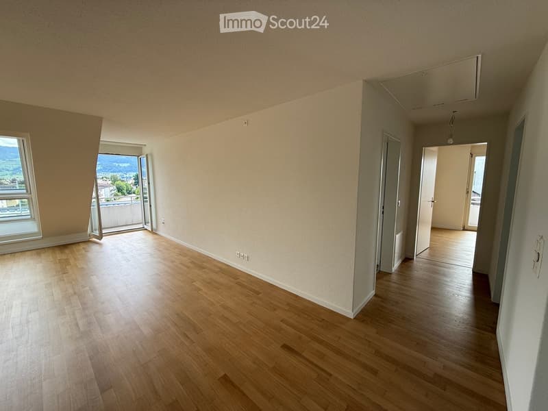 Empty room with wooden flooring, white walls, and large windows offering a scenic view. The room is unfurnished and has an open doorway leading to another room.