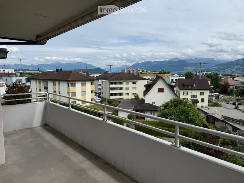 Balcony with white railing, overlooking a residential area with houses, trees, and distant mountains.