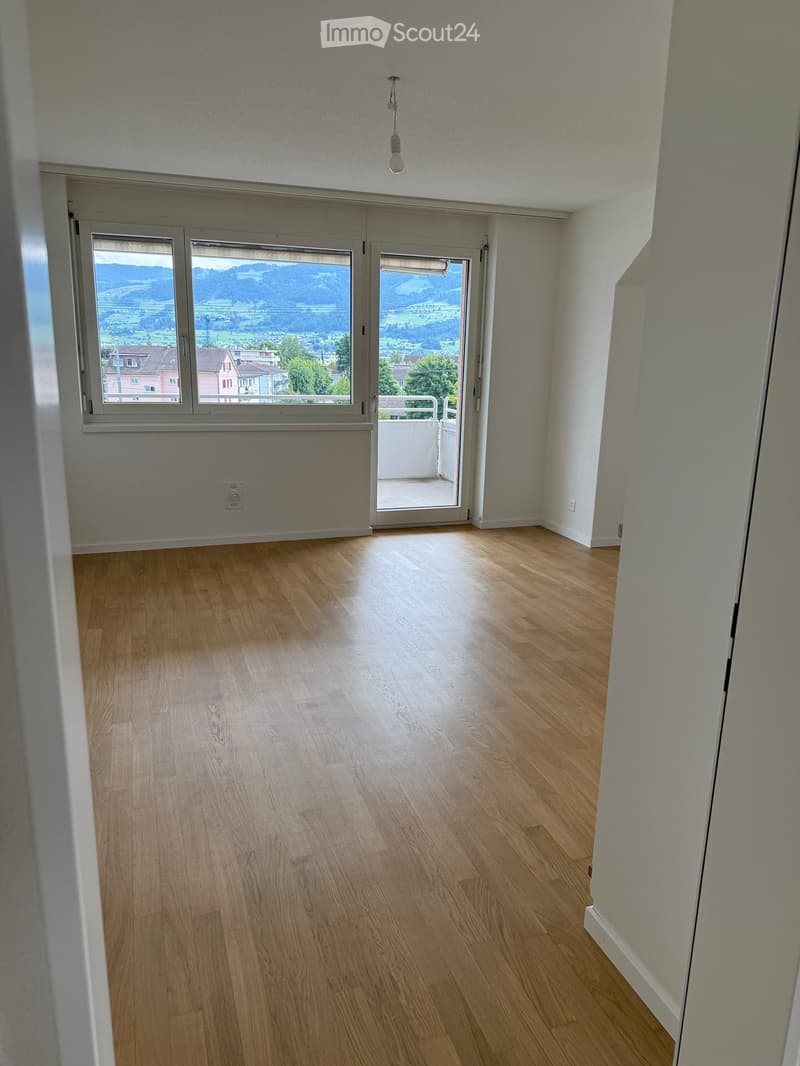 Empty room with wooden flooring, large windows, and a sliding glass door leading to a balcony.