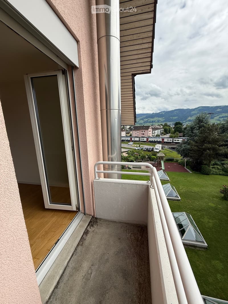 Balcony with wooden floor, white railing, and a view of a green lawn, playground, and distant mountains.