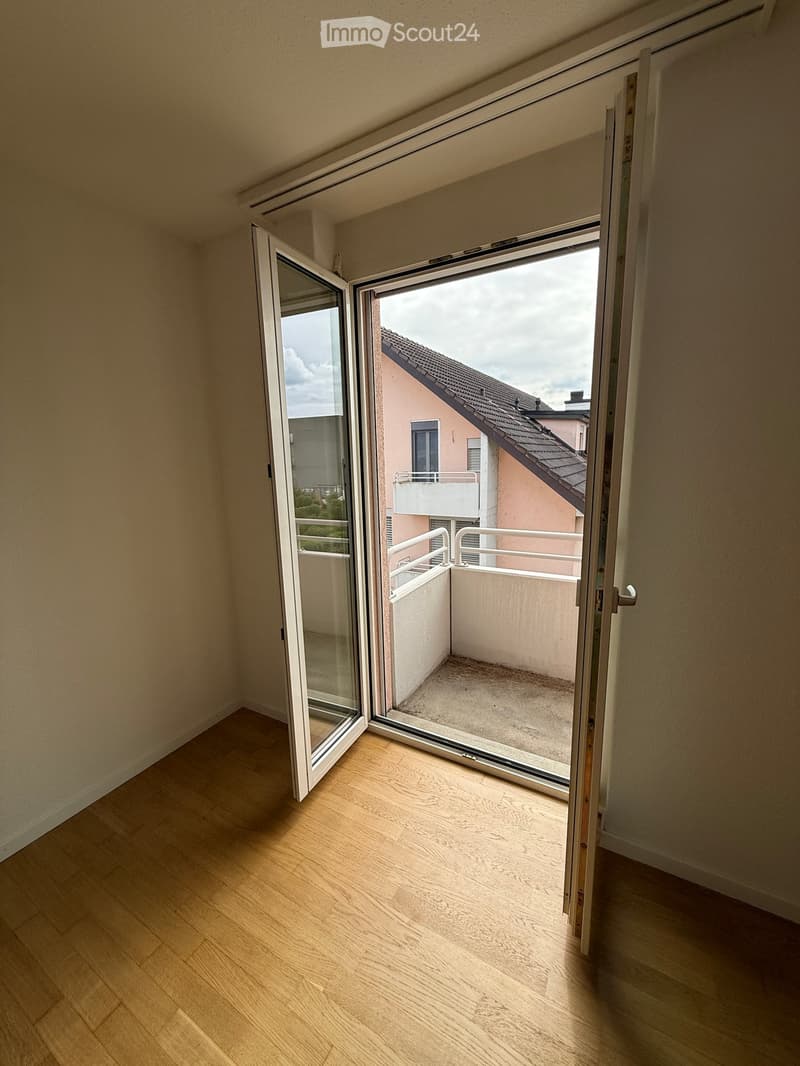 Open sliding glass doors lead to a small balcony with a concrete floor and white railing, overlooking neighboring buildings.