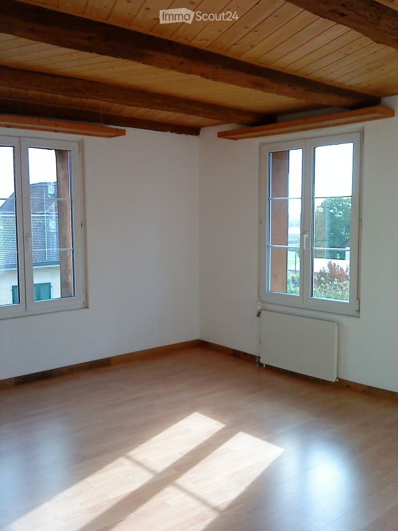 an empty room with wooden floors, white walls, two large windows, a white radiator under the right window, and a wooden ceiling with exposed beams
