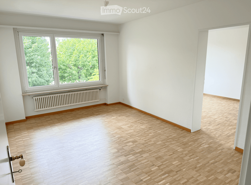 Empty room with wooden flooring, white walls, large window with view of greenery, radiator below window, and a door visible on the left side.